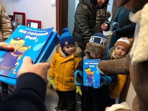 Na pierwszym planie książeczka pt. &quot;Na ratunek Polic&quot; Po prawej policjant, który wręcza taką książeczkę dziecku.