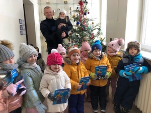 Pamiątkowe zdjęcie, na którym widać policjanta trzymającego dziewczynką na rękach. Przed nimi grupka dzieci, a tle ubrana choinka.