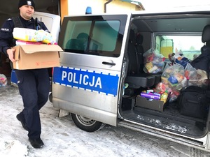 Po lewe policjant, który niesie karton z żywnością. Po prawej stoi radiowóz - bus z otwartymi bocznymi drzwiami.  W samochodzie widoczne paczki z żywnością.
