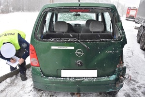 Na zaśnieżonej drodze stoi uszkodzony samochód osobowy. Po lewej policjant wykonujący oględziny auta.