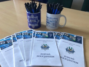 Na stole leżą ulotki z napisem &quot;Zostań policjantem&quot;. Dalej dwa kubeczki, a w nich niebieskie długopisy.