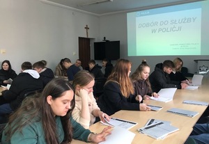 Na pierwszym planie młodzież siedząca przy stołach i  oglądająca jakieś książki. W tle slajd o doborze do służby.