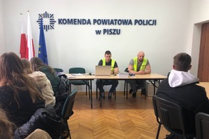 Sala odpraw w komendzie. Na wprost przy stole siedzi dwóch policjantów w kamizelkach odblaskowych. Przed nimi siedzi młodzież słuchająca co mówią policjanci.