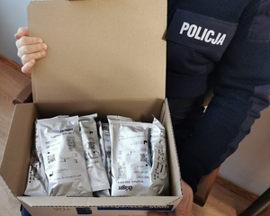 Otwarty karton, w którym widać jakieś opisane opakowania. Karton Trzyma policjant. Widoczny jest fragment jego koszuli z napisem policja.