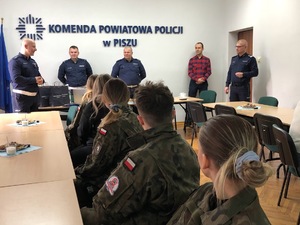 Sala konferencyjna. Przy stołach siedzą panie, a przed nimi stoją policjanci. Jeden z nich przemawia do pań.