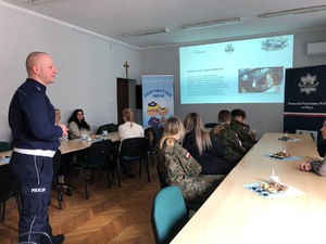 Sala konferencyjna. Przy stołach siedzą panie, które patrzą na wyświetlaną prezentację. Z przodu po lewej policjant, który przemawia do pań.