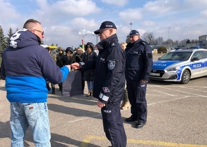 Na placu po prawej policjant, który podaje rękę mężczyźnie stojącemu po lewej. W tle stoją inne osoby oraz radiowóz.