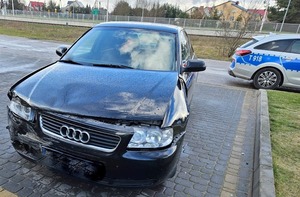 Na parkingu stoi przodem uszkodzone audi. W tle po prawej widać fragment radiowozu.