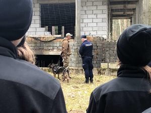 Tyłem stoi dwóch umundurowanych uczniów. Przed nimi policjant i żołnierz z psem.