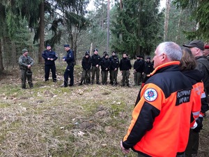 Na polanie w lesie stoją przedstawiciele różnych służb.