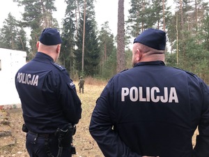 W lesie stoją dwaj policjanci odwróceni plecami.