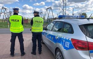Dwaj policjanci piskiej drogówki stojący tyłem. Po ich prawej stronie zaparkowany radiowóz.