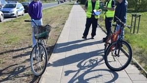 Na pierwszym planie na ścieżce rowerowej stoją dwa rowery. Za nimi policjanci i stojące obok dwie osoby.