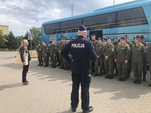 Młodzież ustawiona w dwuszeregu stoi przed autobusem. Przed nimi stoi policjant.