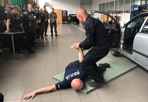 Młodzież na sali gimnastycznej. Przed nimi na materacu leży policjant, a drugi trzyma go za rękę.