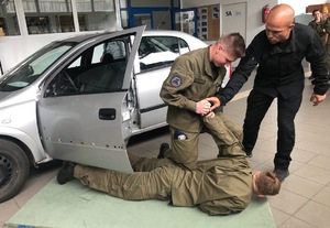 Samochód, a przed nim na materacu leży uczeń. Drugi trzyma go za rękę. Po prawej stoi policjant.