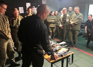 Młodzież na strzelnicy. Przed nimi stoi instruktor, który prezentuje broń.