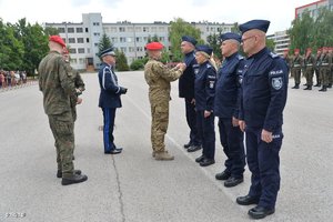 Na placu stoi kilku policjantów. Naprzeciwko nich policjant jednemu z nich podaje rękę.
