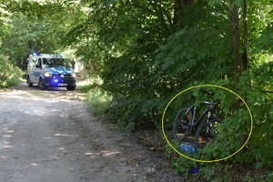 Droga leśna. Po lewej w krzakach rower. W tle na drodze stoi policyjny bus.