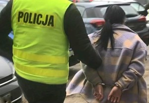 Policjant w kamizelce odblaskowej prowadzi kobietę w kajdankach.